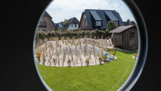 Vista a través de un ventanal circular que muestra una zona residencial con casas, una estructura de barril de madera y un césped.