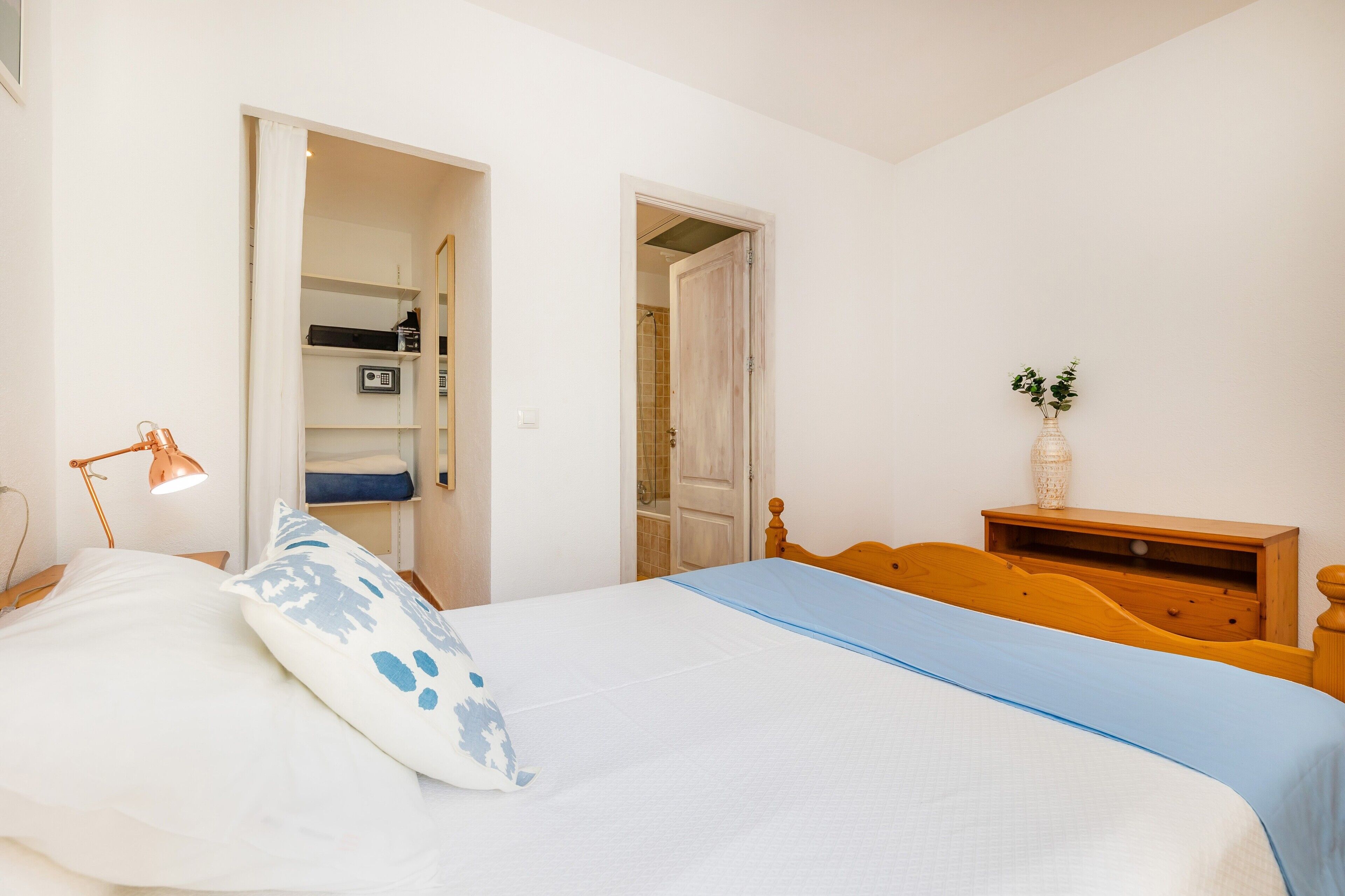 A bedroom with a bed, a nightstand, a lamp, a dresser, and a door leading to a bathroom.