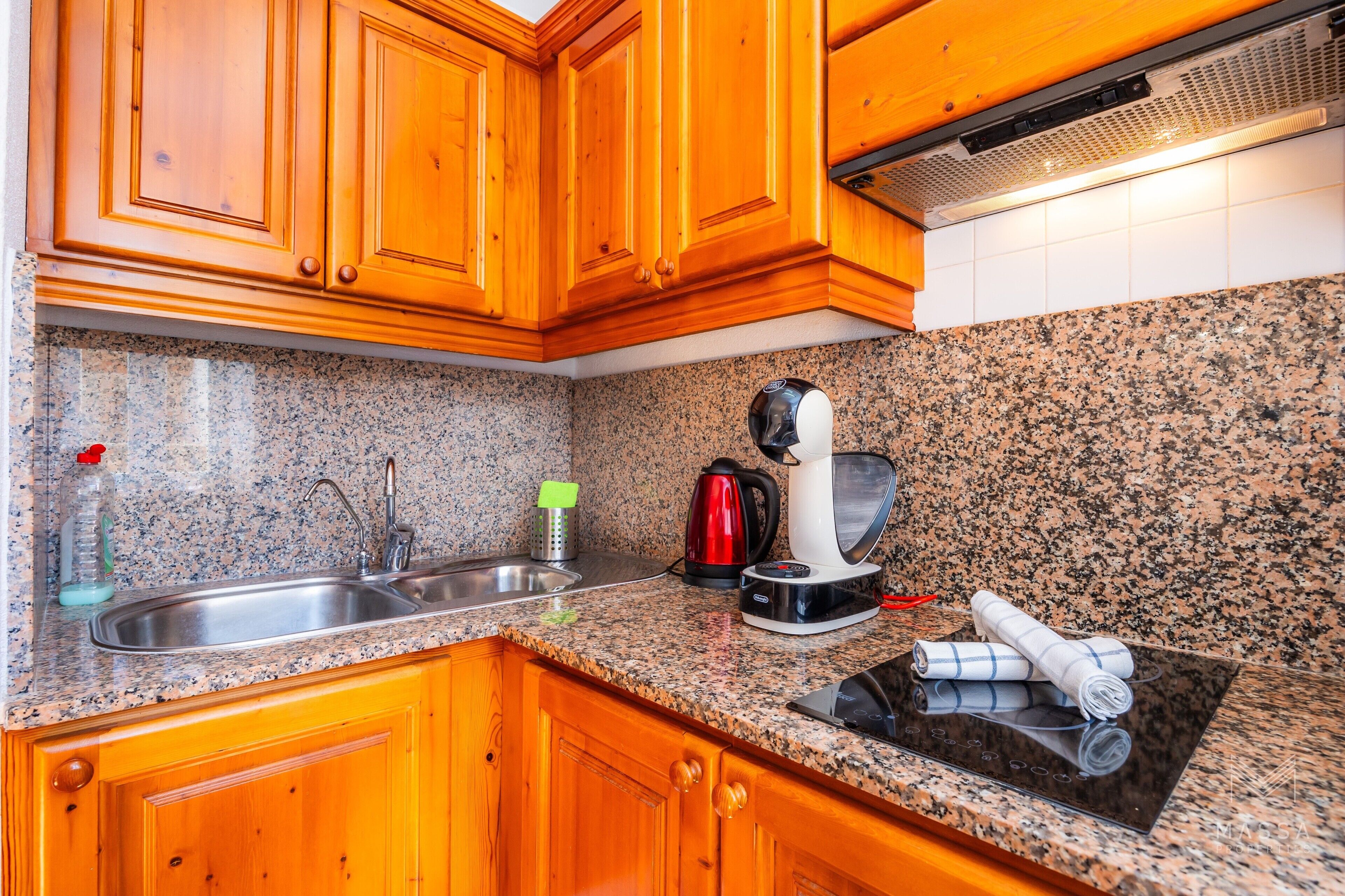 A kitchen with granite countertops, wooden cabinets, a stainless steel sink, a red kettle, a coffee maker, and a black induction cooktop.