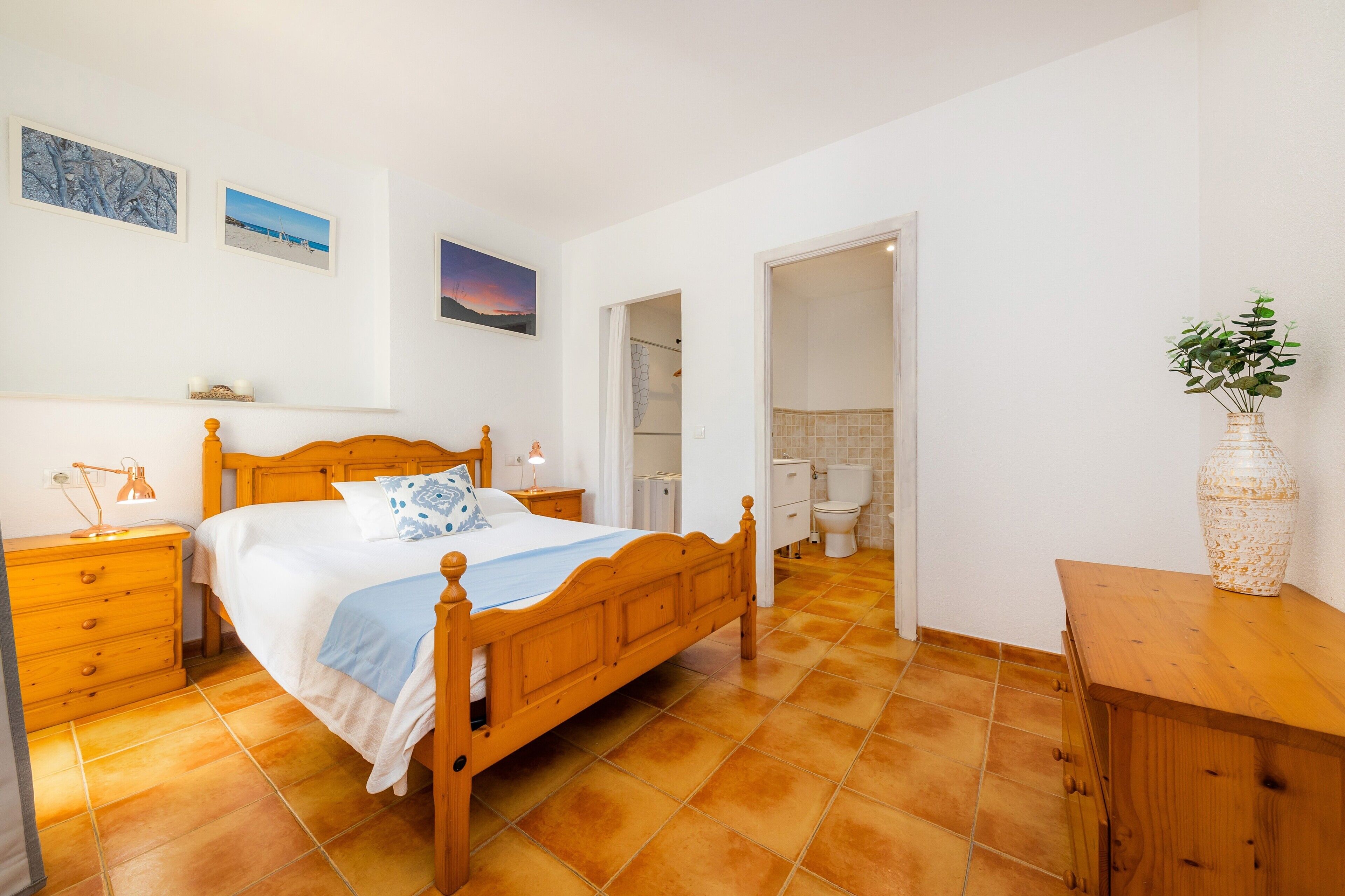 Ein Schlafzimmer mit einem hölzernen Bett, einem Nachttisch, einer Kommode, einem durchgegangenen Tür erkennbaren Badezimmer und einer Vase mit Pflanzen auf einem Beistelltisch.