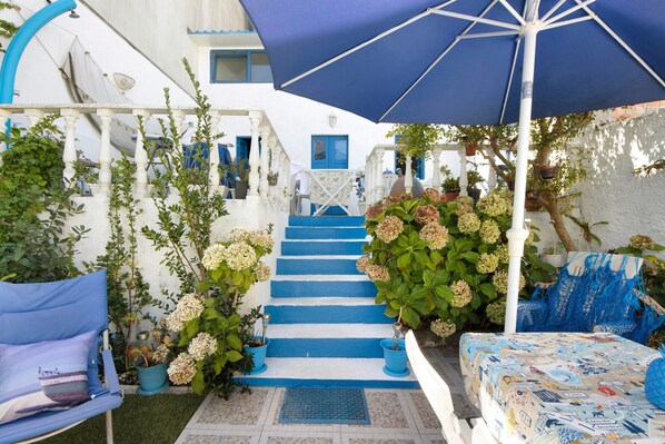 Un bâtiment blanc agrémenté de touches de bleu, un escalier, un parapluie bleu et des plantes en pot.
