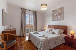 Ein Schlafzimmer mit Bett, Nachttischlampen, einem Sessel, einem Schreibtisch, einem Gemälde und einem Fenster mit Vorhängen.