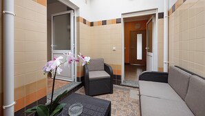 Ein überdachter Außenbereich mit Korbmöbeln, einem Sessel und einer Couch, einer Topf-Orchidee und einem Fliesenboden.