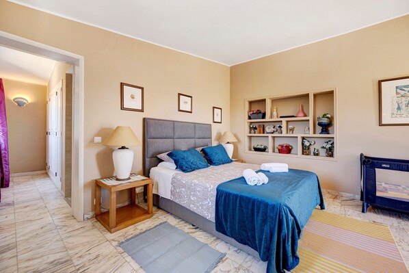 A hotel room with a bed, bedside lamps, a nightstand, a shelf with decorative items, and a door leading to another room.