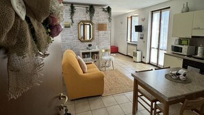 Un salon moderne avec un canapé jaune, une table à manger avec des chaises, un coin cuisine avec des placards et des appareils électroménagers, et un mur en pierre agrémenté de plantes.