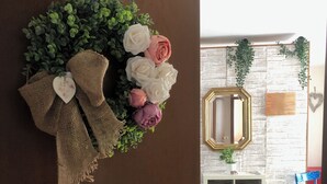 Une pièce dont le mur est décoré dans un style rustique, avec du tissu de jute et des fleurs, une bibliothèque blanche remplie de livres et un fauteuil bleu.