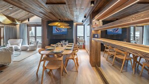 A spacious dining area with a long wooden table and chairs, a bar area with a wooden counter, and a large window offering a view of the outdoors. - New apartment in Méribel with sauna and parking (Méribel)