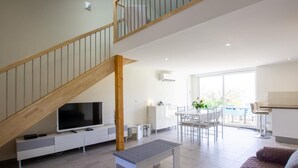 A modern living room with a wooden staircase, a flat-screen TV, a dining area, and a large window.