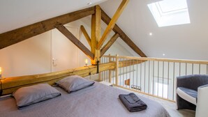 A bedroom with a sloped ceiling, wooden beams, a bed with pillows, a chair, and a skylight.