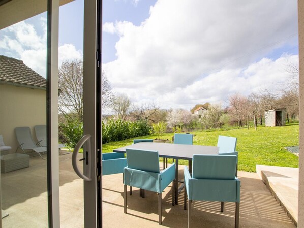 A patio with a table and chairs, a glass door, and a view of a grassy area with trees and a building in the background.