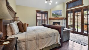 A bedroom with a bed, bedside table, lamp, fireplace, and large windows.
