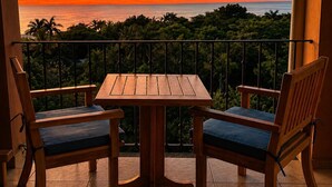 Een balkon met een houten tafel en twee stoelen, met uitzicht op een weelderig landschap en een zonsondergang.