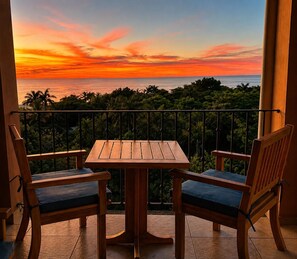 A balcony with a wooden table and two chairs, overlooking a lush landscape and a sunset.