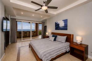 Un dormitorio con cama, ventilador de techo, televisión, balcón con vistas y un cuadro en la pared.