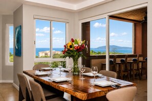 A dining room with a wooden table set for six, large windows with ocean views, and a vase of flowers on the table.