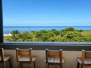 A balcony with three wooden chairs and a view of the ocean and tropical foliage.