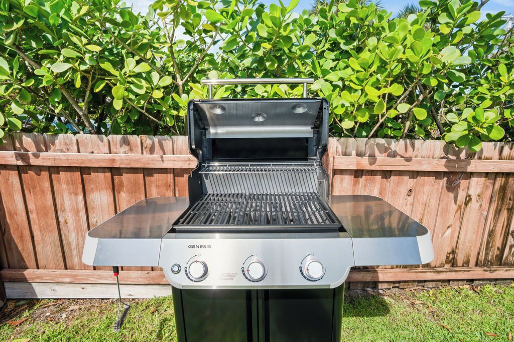 Ein Genesis tragbarer Gasgrill mit digitaler Anzeige, einem Seitenbrenner und einer Ablagehaube, vor einer Holzlattenverkleidung und grünem Laub.