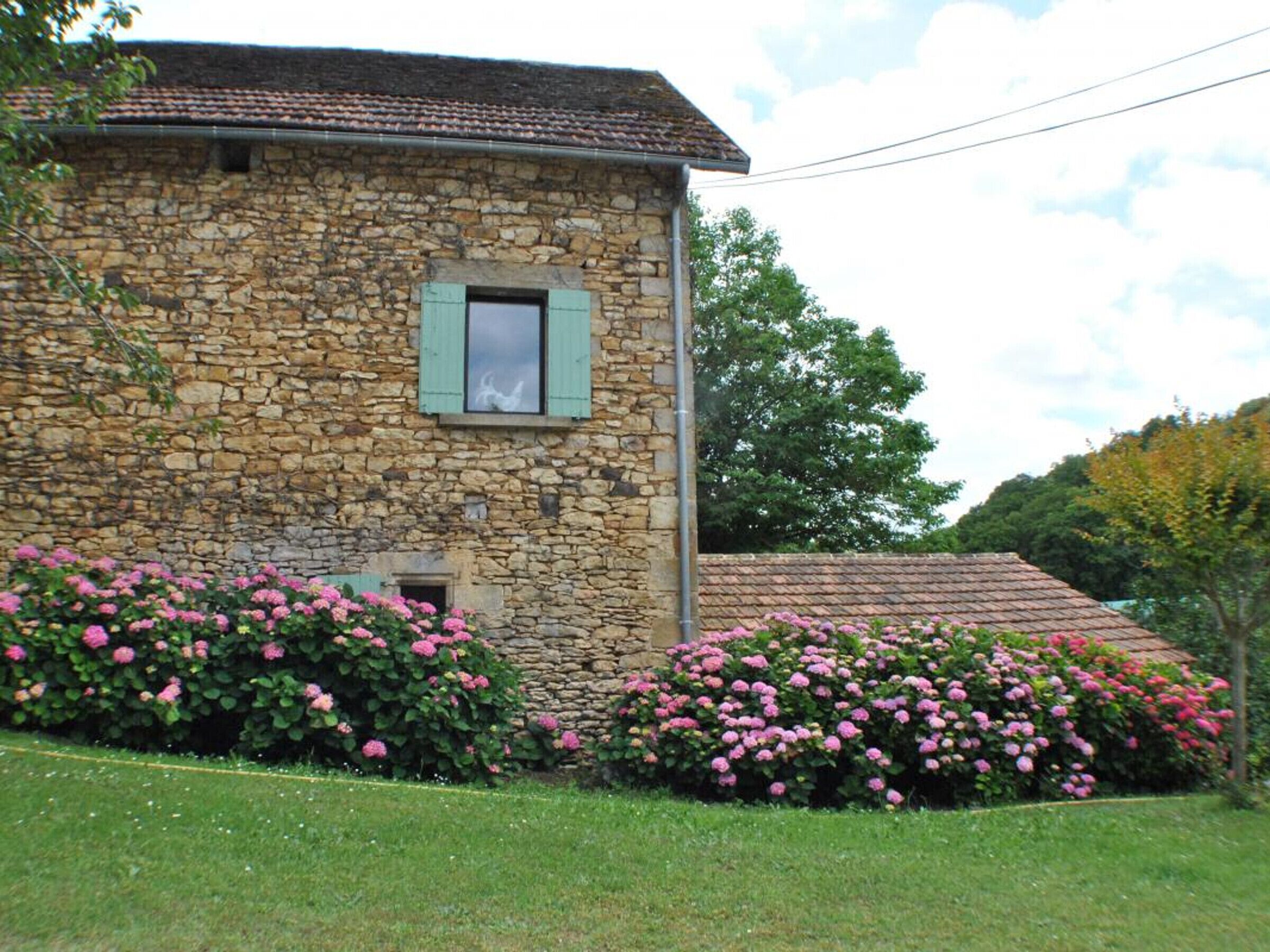Un bâtiment en pierre avec une petite fenêtre et une porte, entouré d’arbustes à fleurs roses.