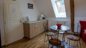 Een kleine keuken met houten kastjes, een wit aanrecht, een magnetron en een koffiezetapparaat. Er staat een eettafel met metalen stoelen en een rode kom. Een klok hangt aan de muur, en een raam zorgt voor natuurlijk licht.