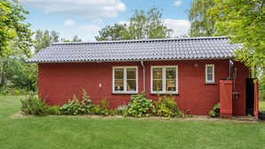 A red house with a grey tiled roof, two windows, and a small garden area with green grass and plants.