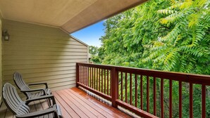 A wooden deck with two Adirondack chairs, a railing, and a view of dense green foliage.