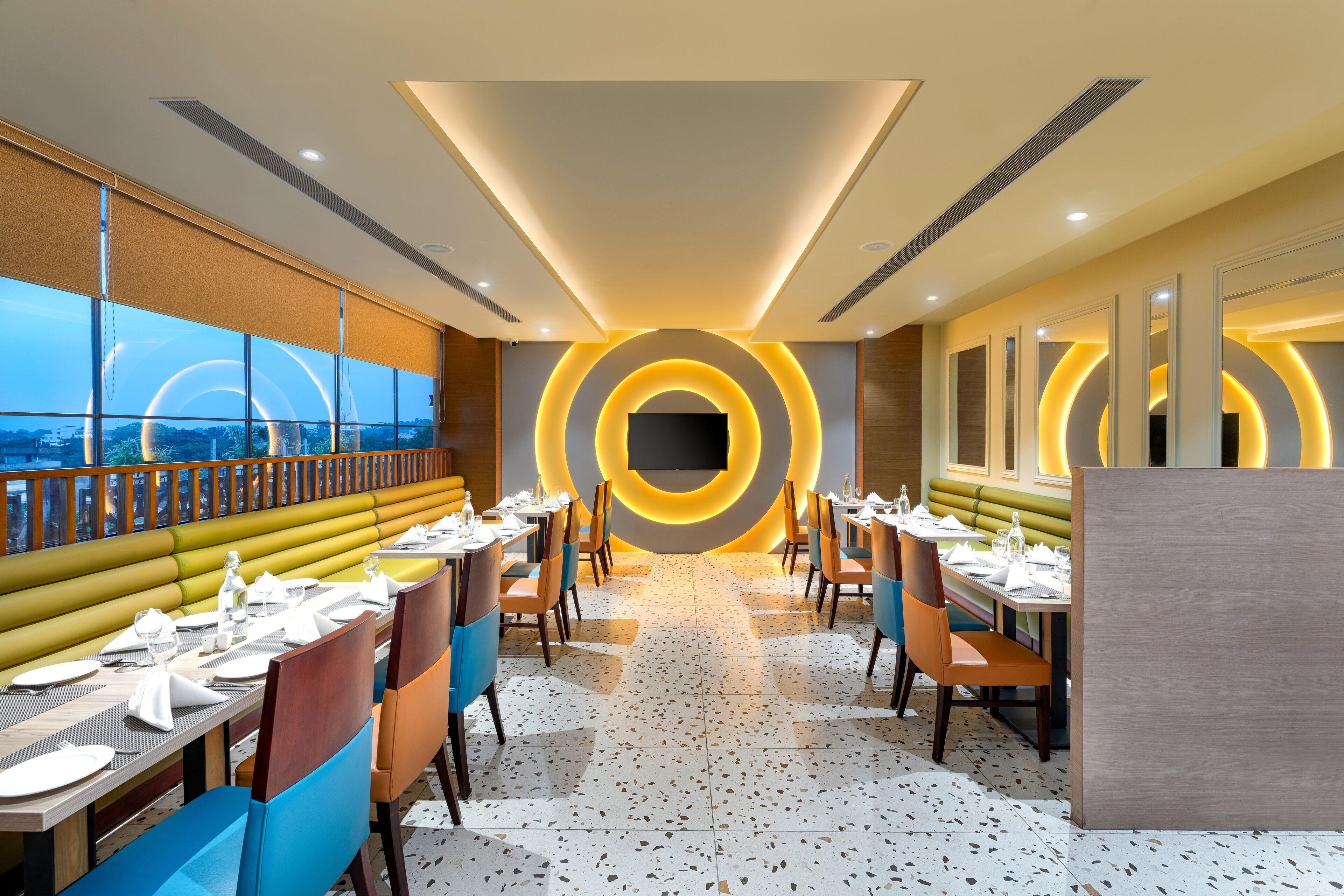 A modern restaurant with a central circular light feature, long tables with chairs, and a view through large windows.