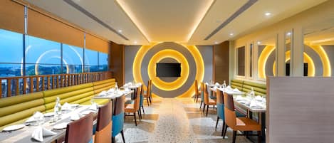 A modern restaurant with a central circular light feature, long tables with chairs, and a view through large windows.