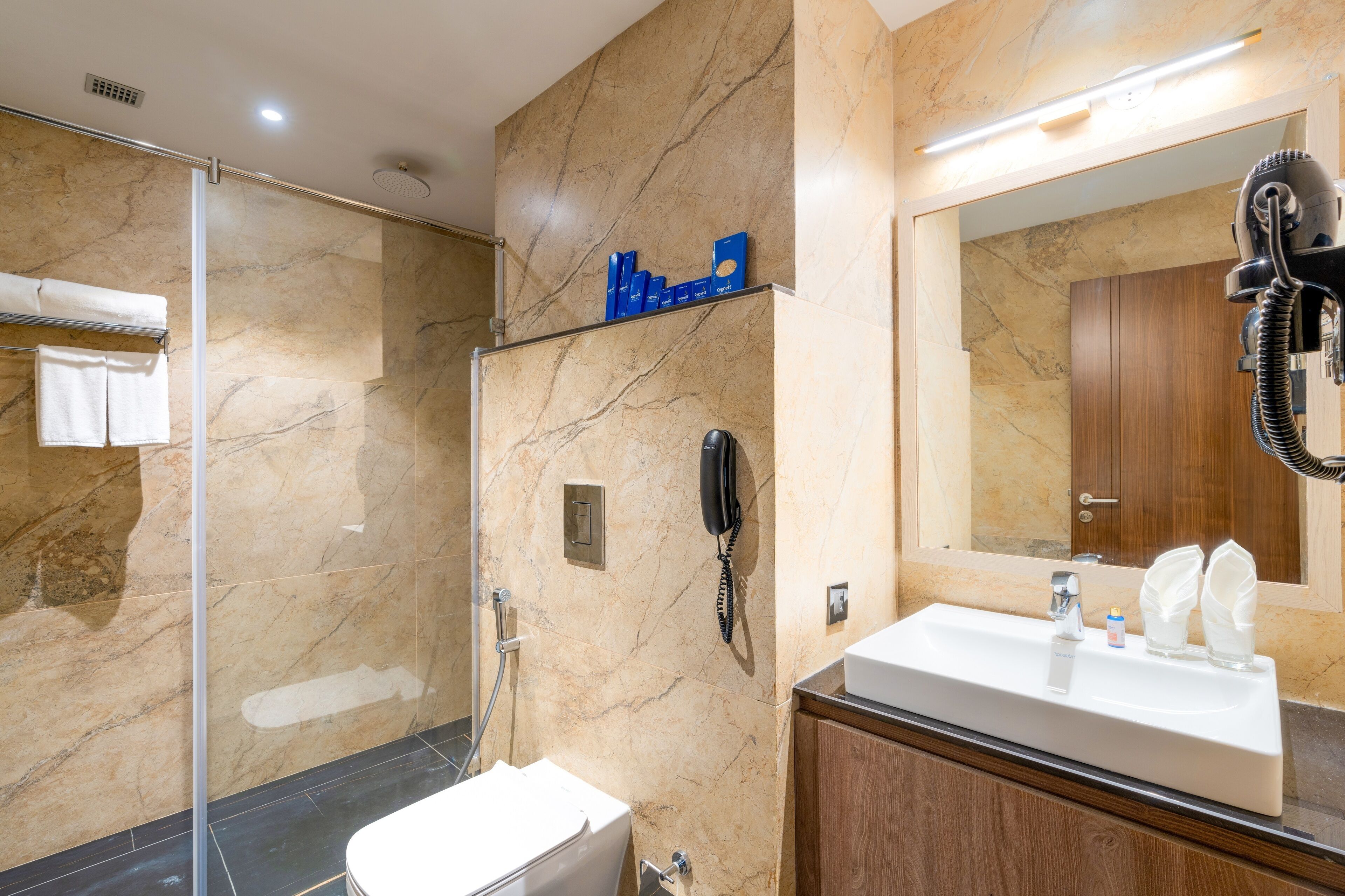 A bathroom with a marble wall, a glass shower enclosure, a white sink, and a wooden vanity.