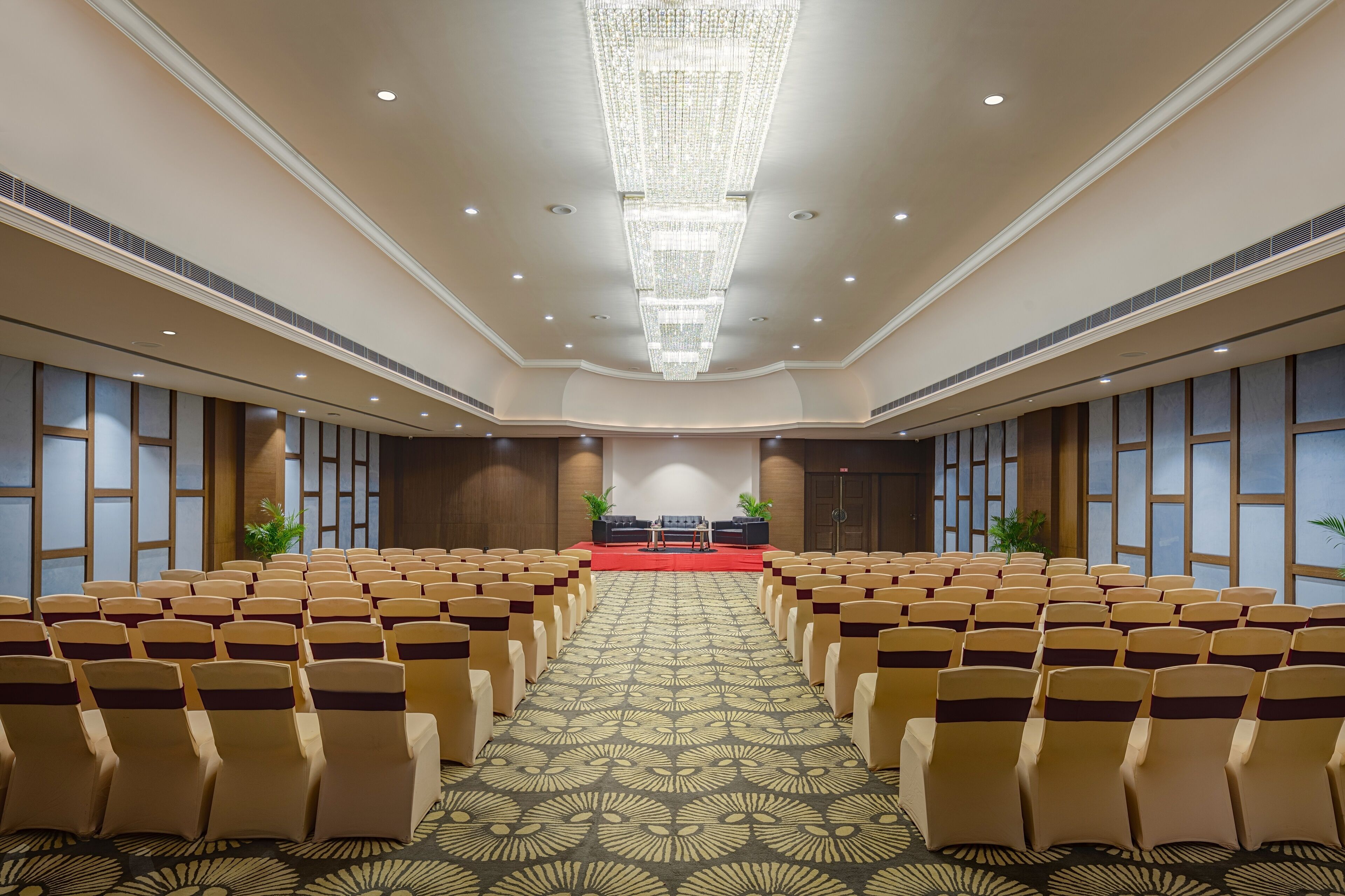Uma sala de conferências ampla com fileiras de cadeiras, palco com carpete vermelho e um candelabro.