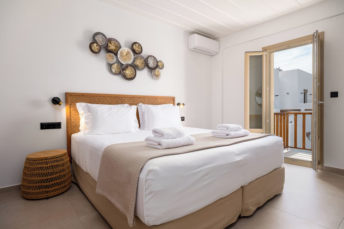 Un lit bien fait, avec du linge de lit blanc et un plaid beige, un tabouret rond en osier et un élément décoratif fixé au mur.