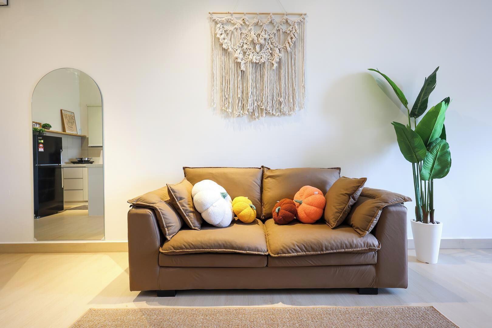 A brown leather sofa with multiple cushions, a macrame wall hanging, a potted plant, and a kitchen area visible through an archway.
