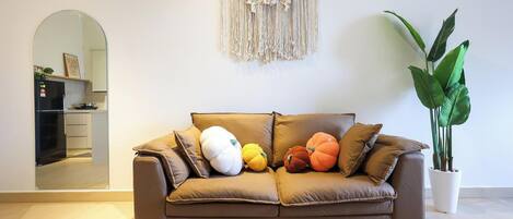 A brown leather sofa with multiple cushions, a macrame wall hanging, a potted plant, and a kitchen area visible through an archway.