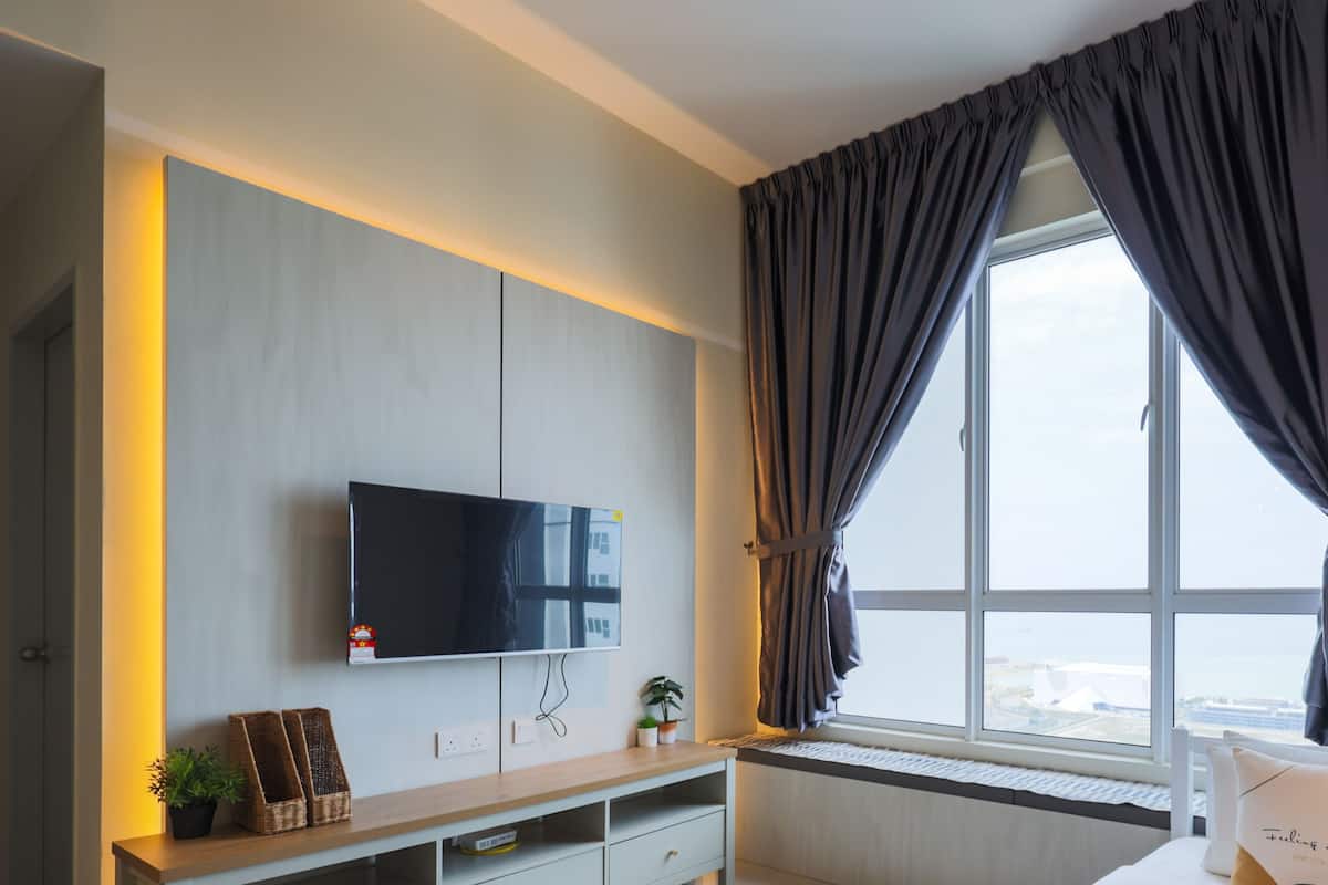 A modern hotel room with a large flat-screen TV mounted on the wall, a window with dark curtains, a small plant on the windowsill, and a bedside table with a pillow.