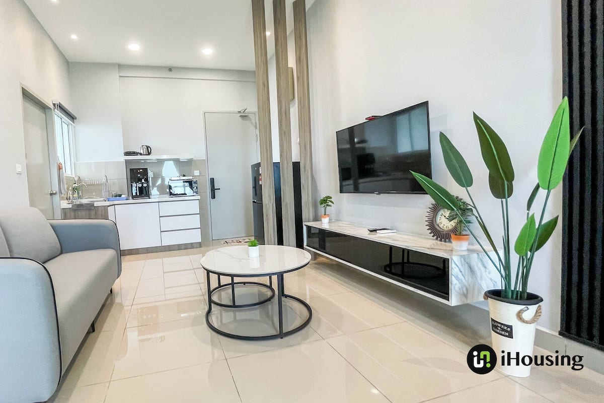 A modern living room with a grey sofa, a round coffee table, a flat-screen TV, and a kitchen in the background.