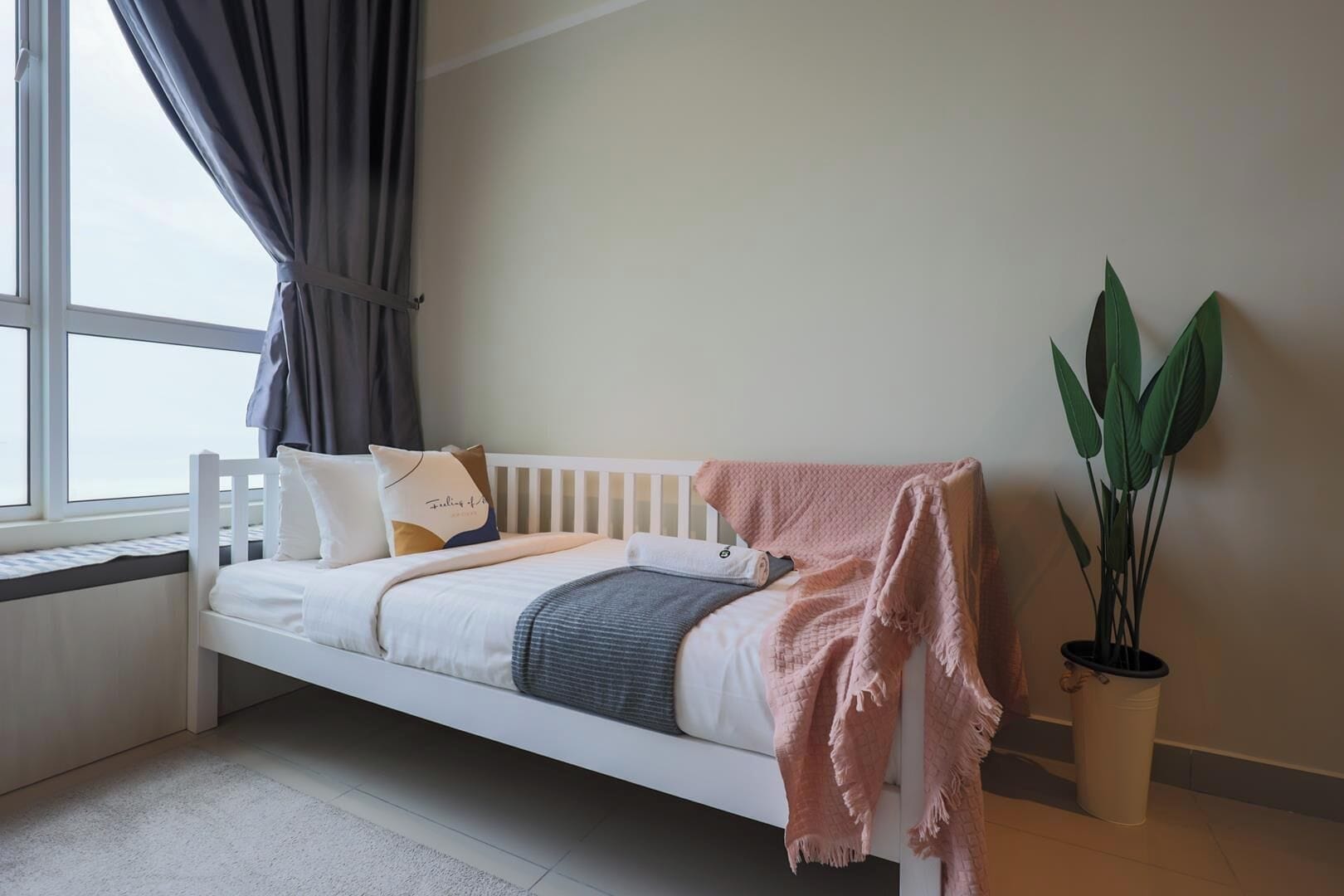 Una habitación con cama blanca, ventana con cortinas, planta en maceta y radiador.