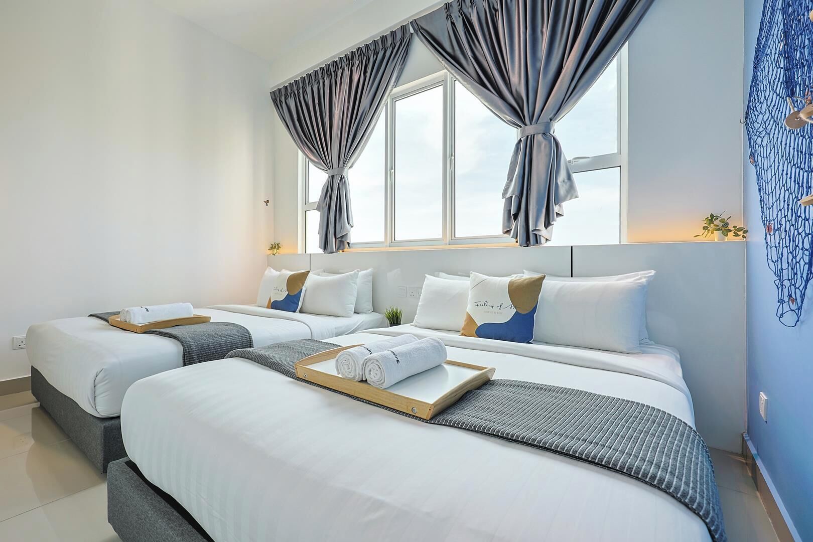 A hotel room with two beds, a large window with curtains, a bedside table with towels, and a wall-mounted plant.