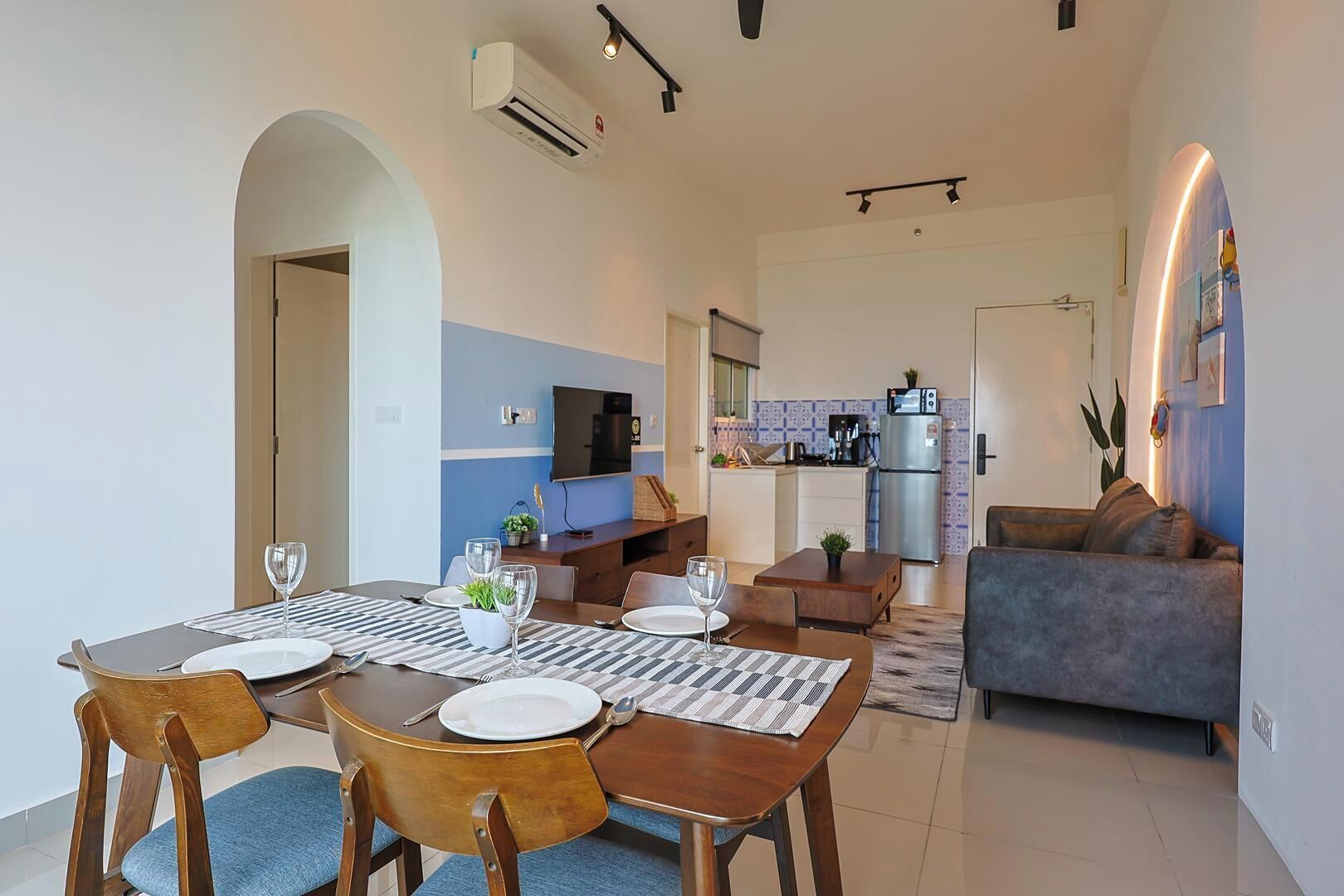 Uma sala de estar moderna com um sofá, uma área de jantar com mesa posta para quatro pessoas e uma cozinha ao fundo.