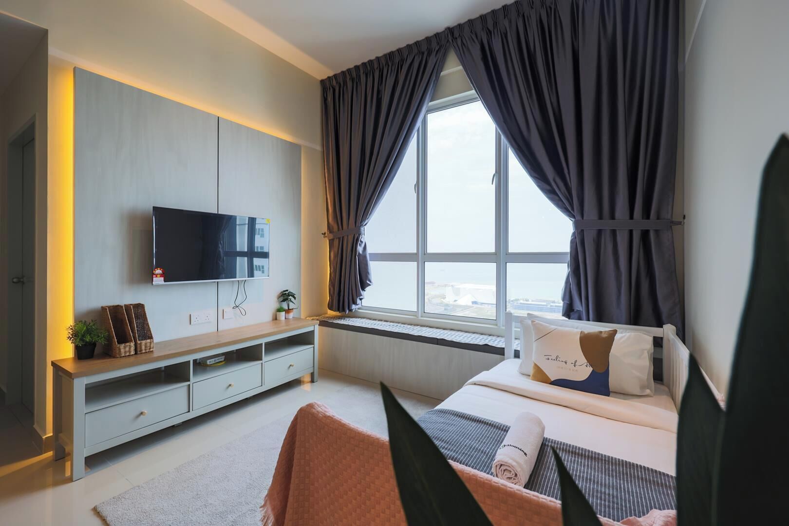 Ein modernes Schlafzimmer mit Wand-Fernseher, Nachttisch, Bett mit Kissen und einem Fenster mit Vorhängen sowie Ausblick.