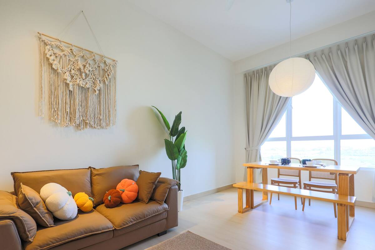 A modern living room with a brown leather sofa, a macrame wall hanging, a large potted plant, a wooden dining table with chairs, and a large window with curtains.