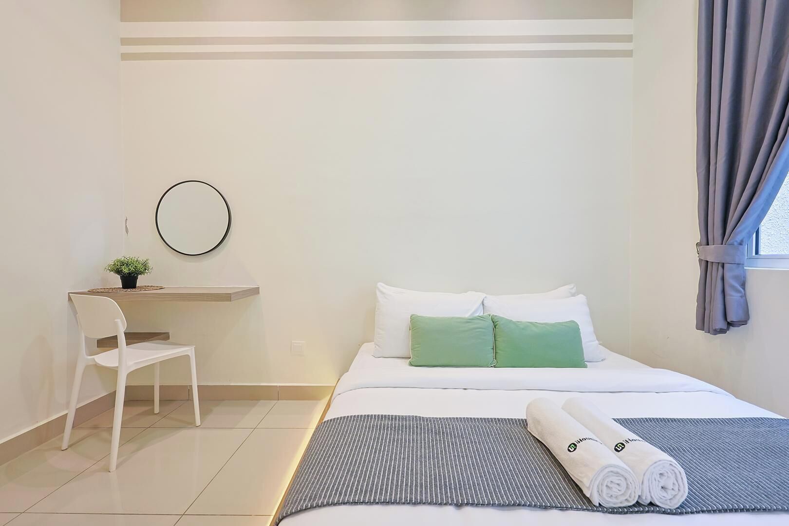 A hotel room with a bed, a desk with a chair, a small plant, and a window with curtains.