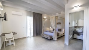 A modern bedroom with a four-poster bed, a bedside table, a chair, and a bathroom with a sink and mirror. - PietraBianca Marzamemi (Syracuse)