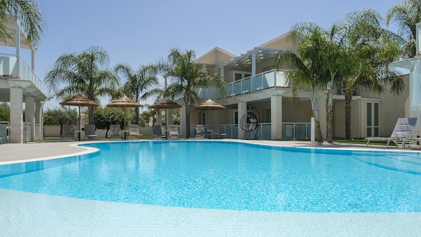 A swimming pool with lounge chairs and umbrellas, surrounded by palm trees and a multi-story building. - PietraBianca Marzamemi (Syracuse)