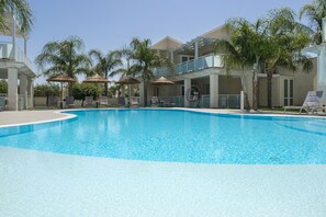 A swimming pool with lounge chairs and umbrellas, surrounded by palm trees and a multi-story building. - PietraBianca Marzamemi (Syracuse)