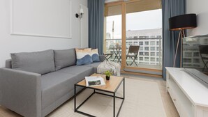 A modern living room with a grey sofa, a wooden coffee table, a TV, and a balcony with a view of buildings.