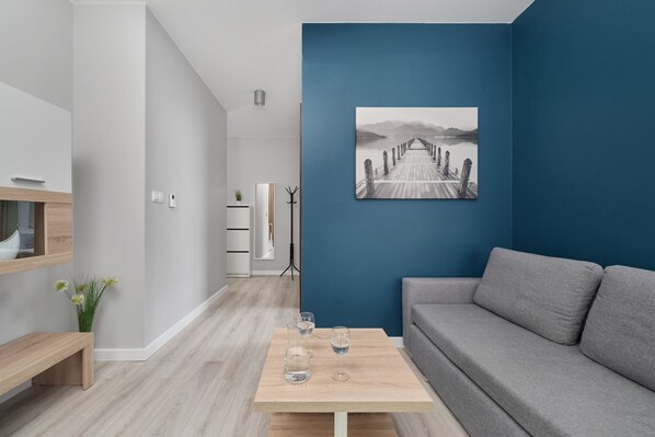 A modern living room with a grey sofa, a wooden coffee table with glasses, a blue accent wall, and a framed picture of a pier.