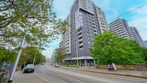 A modern high-rise building with multiple floors, a pedestrian walkway, and a tram line. - Apartment with River View | Balcony and Parking (Wrocław)