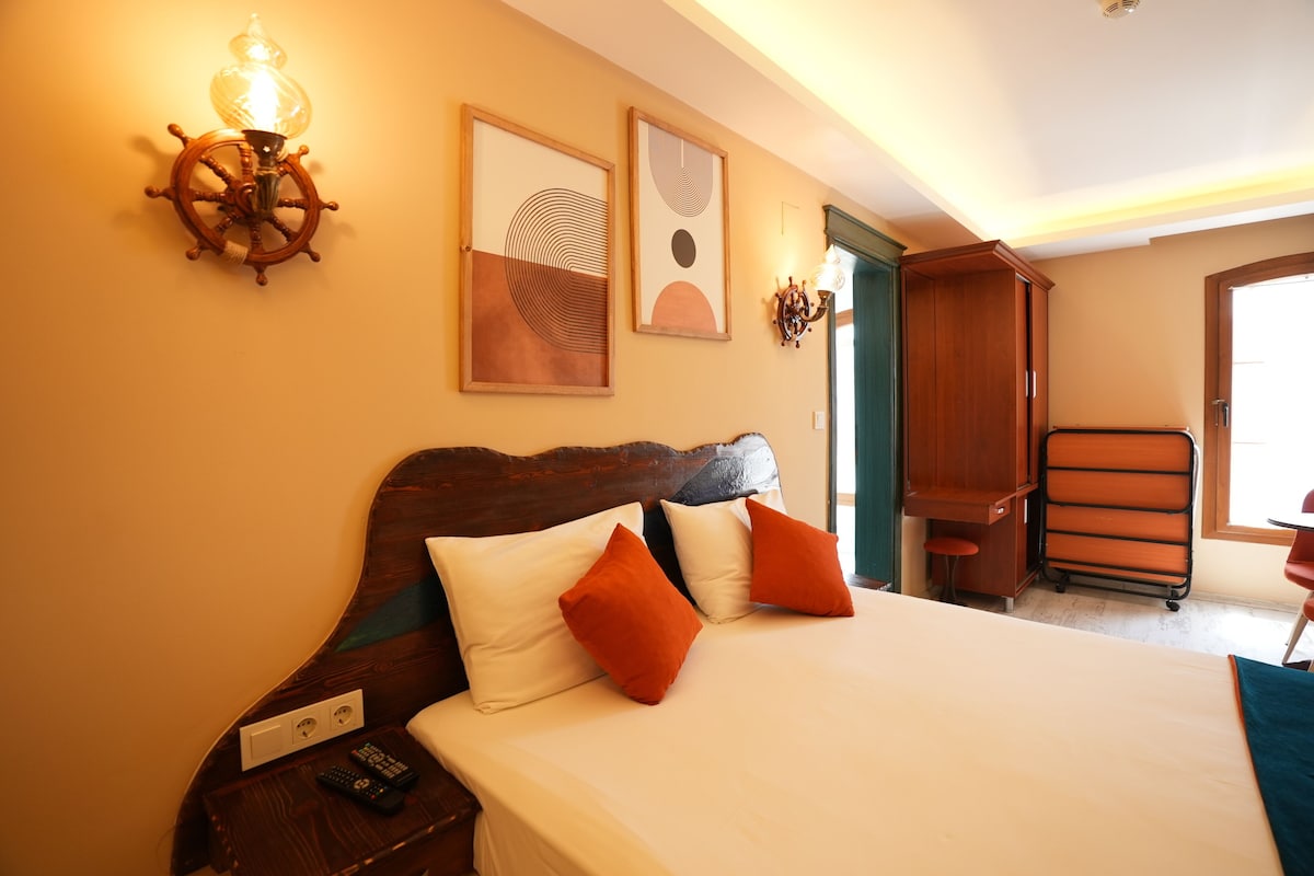 Habitación de hotel con cabecera de madera, cama con almohadas blancas y anaranjadas, mesita de noche con control remoto y obras de arte en las paredes.