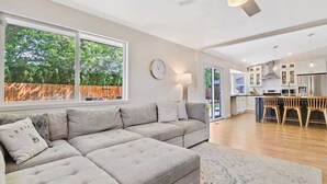 A modern living room with a sectional sofa, a dining area with wooden chairs, a kitchen with white cabinets, and a large window with a view of trees.