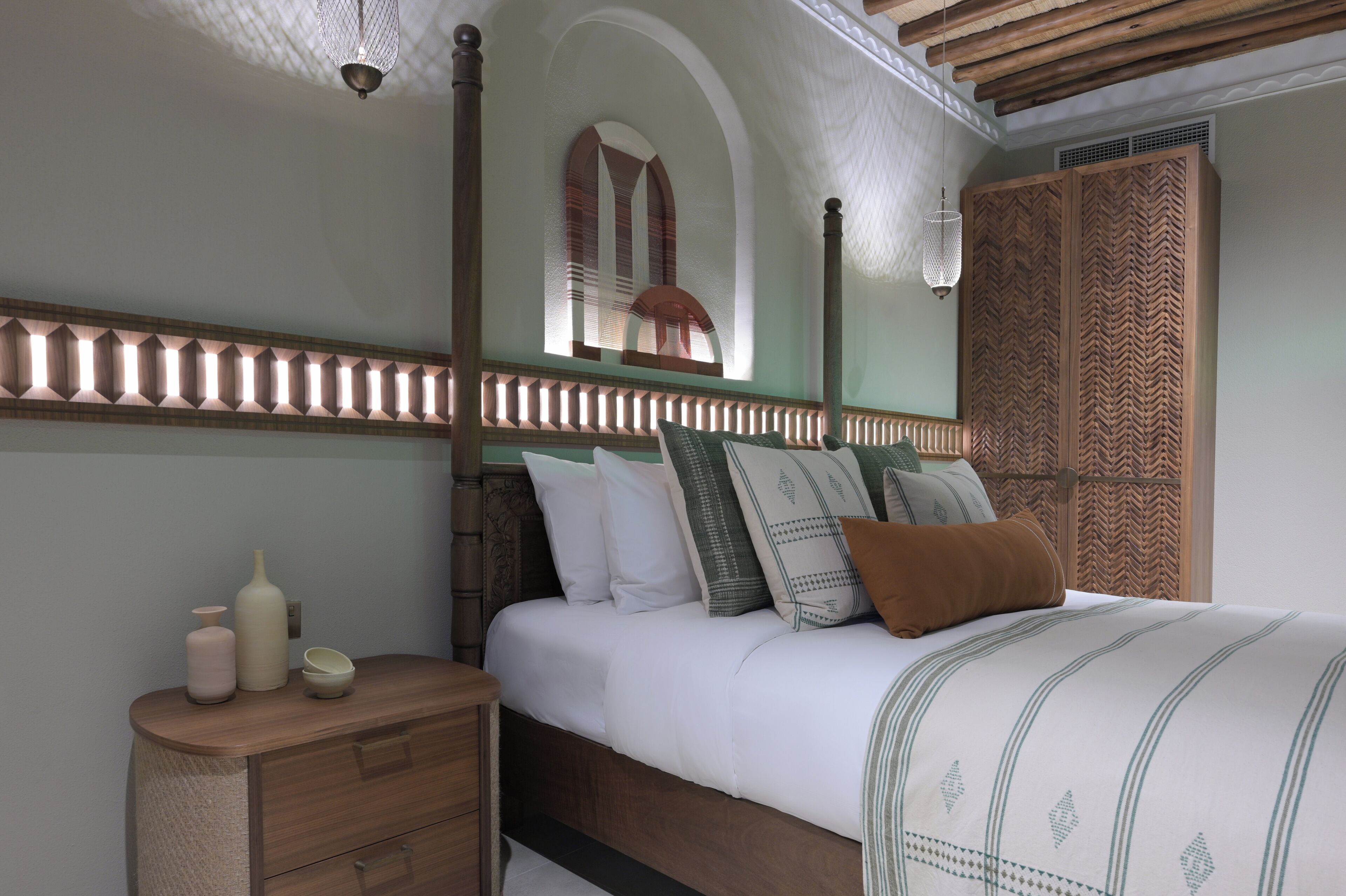 A bedroom with a wooden headboard, a bed with white bedding, a nightstand with decorative items, and a large window with a view of the outside.