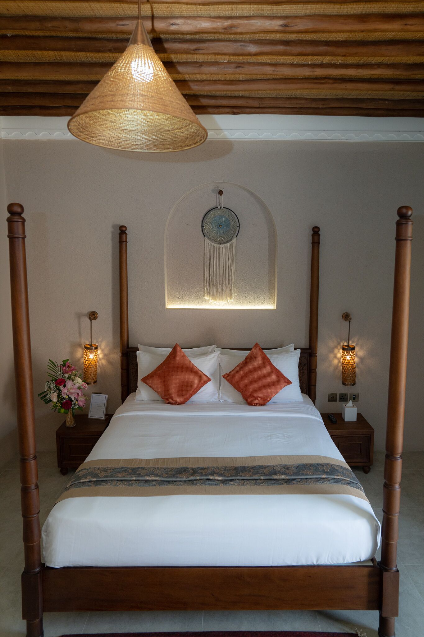 Uma cama de dossel com colcha branca e estampada, dois travesseiros laranja e cabeceira de madeira. Há duas mesas de cabeceira com abajures e um vaso de flores.
